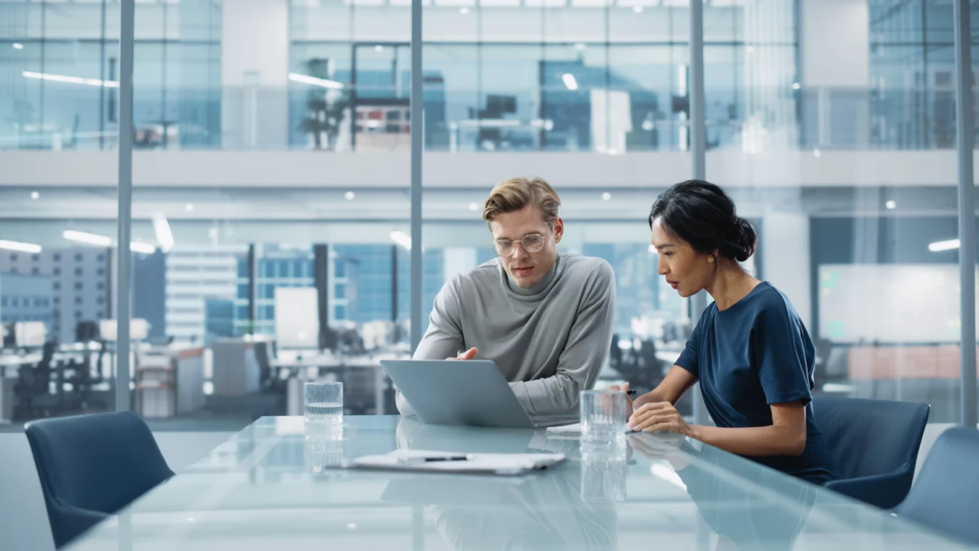 A man and a woman looking at a laptop together at a glass conference table in a modern office.