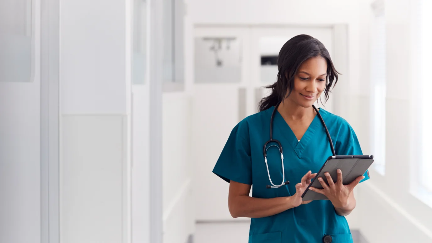 A healthcare worker in scrubs and a stethoscope walking in a hospital hall while looking at a tablet.