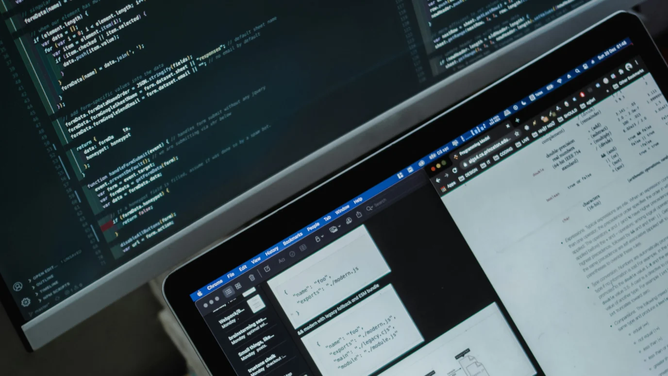 Close-up of a desktop monitor showing lines of code (JavaScript/React) next to a laptop displaying technical documentation and JSON data, emphasizing advanced coding and data strategy in an accelerator.
