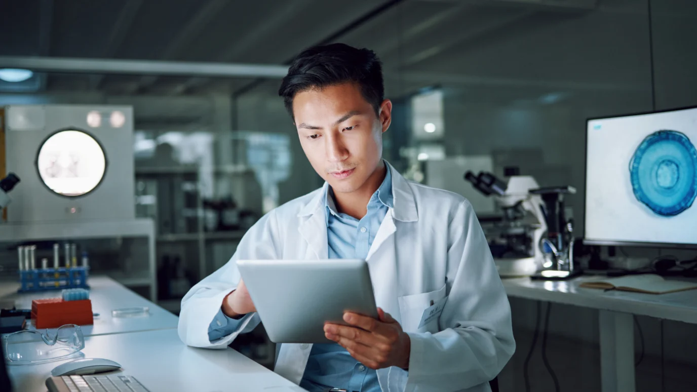 Focused scientist or data analyst in a white lab coat using a digital tablet, with a microscope and scientific data displayed on a monitor, emphasizing AI-driven discovery and data analysis in a laboratory setting.