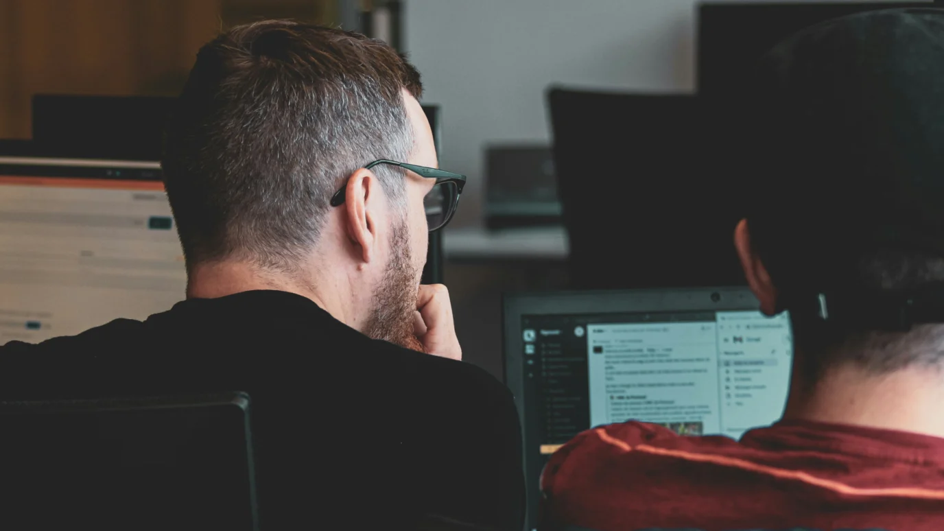 Rear view of two male founders or engineers reviewing code, infrastructure, or a software platform on a laptop screen, emphasizing the hands-on technical guidance available in an accelerator.