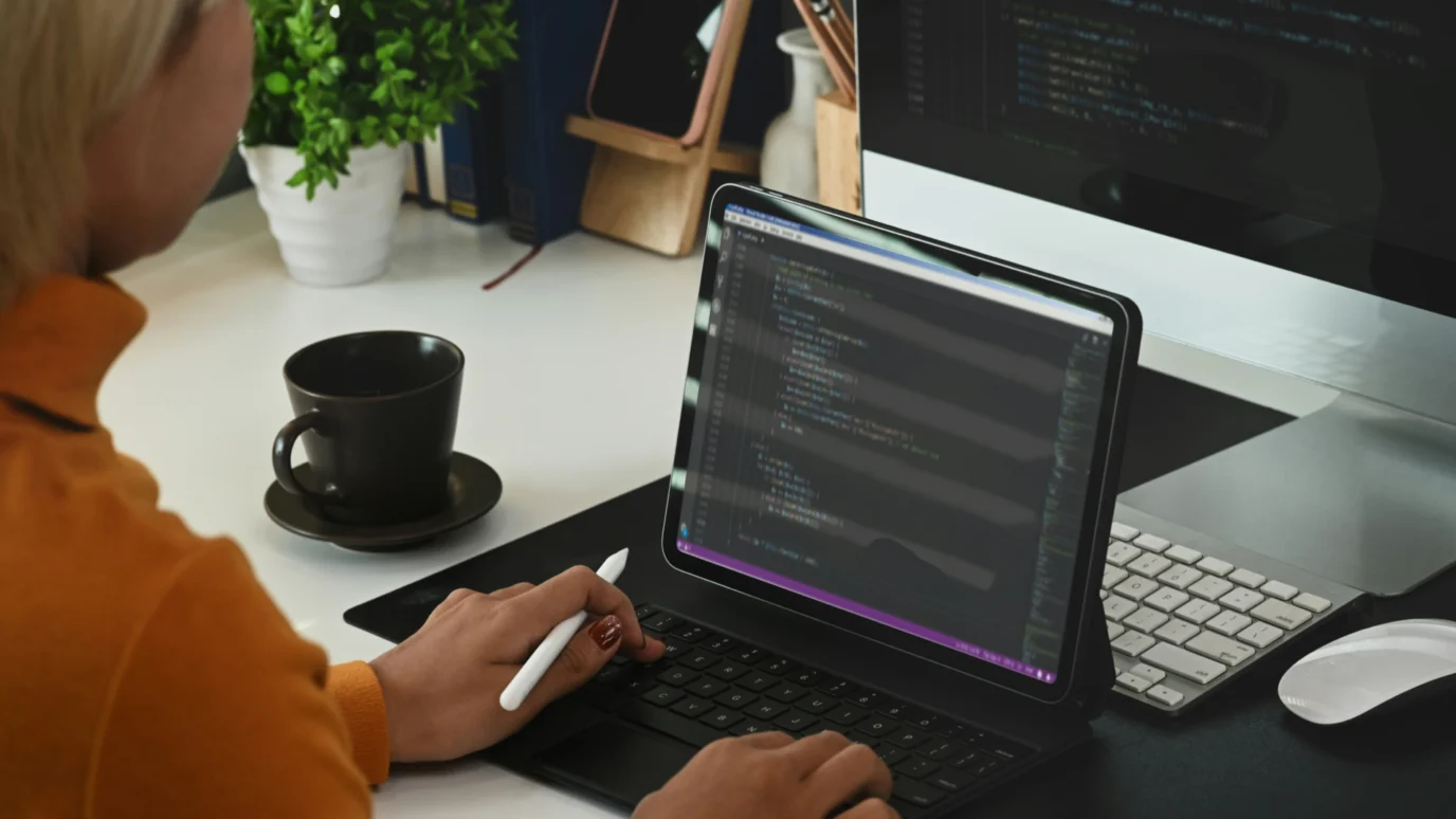 Woman coder or engineer using a stylus and tablet keyboard, with a desktop monitor displaying complex software code in the background, emphasizing bespoke solutions and technical expertise.
