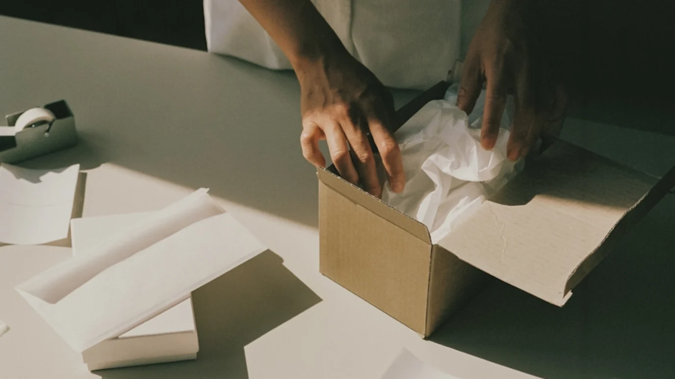 Close-up of hands carefully packing a product into a cardboard shipping box with white tissue paper, emphasizing quality control, branding, and customer experience for a scalable startup.