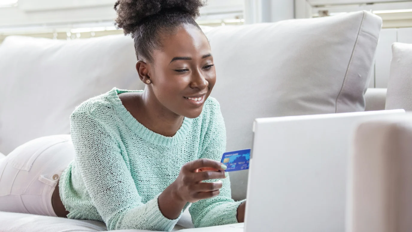Happy young African-American woman holding a blue credit card and smiling while shopping online on a laptop, emphasizing user trust and checkout conversion for an accelerated startup.