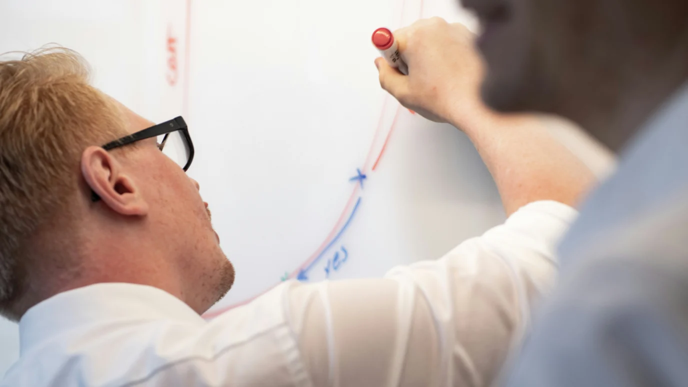 Person with glasses drawing a diagram on a whiteboard with a red marker.