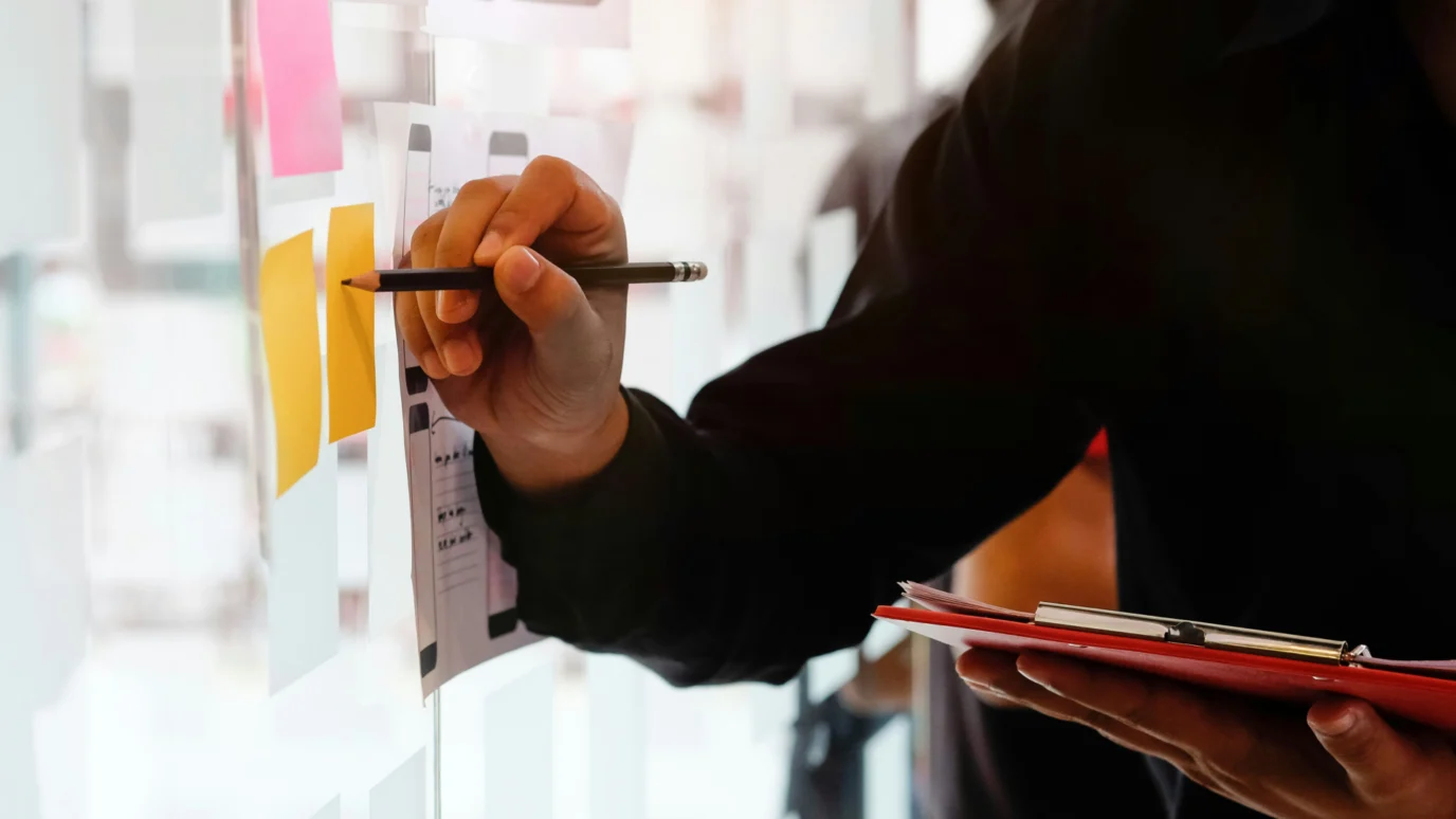 Hand writing on a paper wireframe attached to a glass wall with sticky notes.
