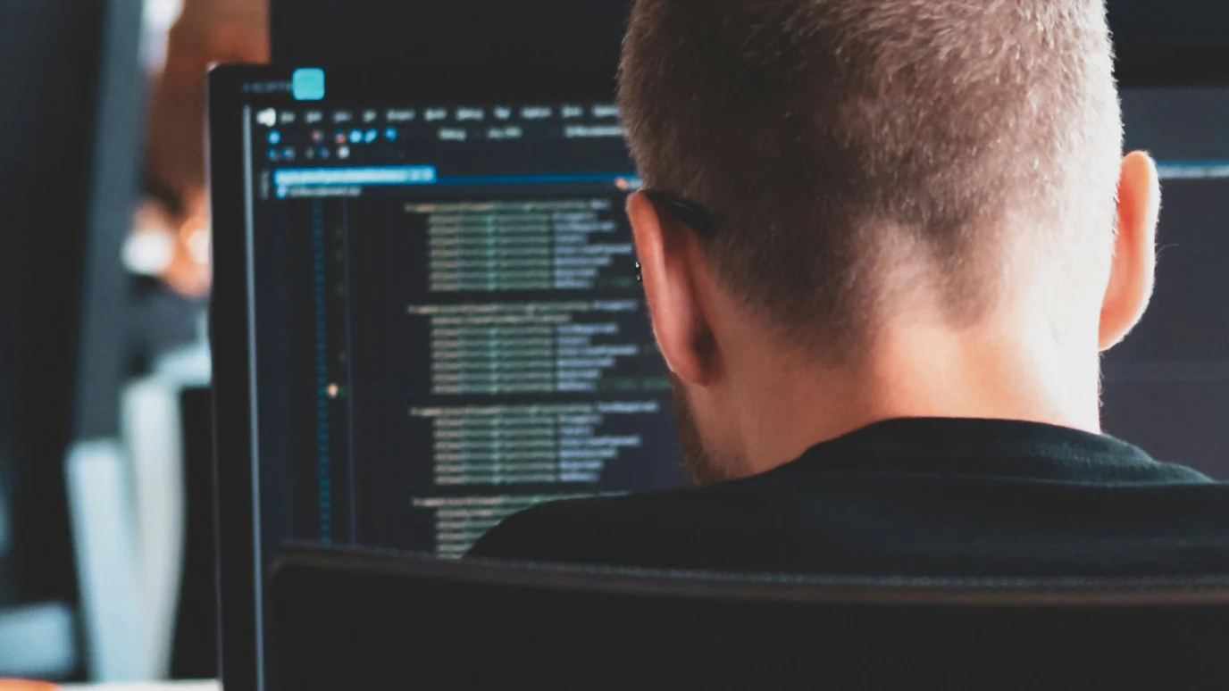 Close-up rear view of a focused engineer or developer reviewing lines of code on a desktop monitor, emphasizing back-end technology, quality assurance (QA), and deep technical work in an accelerator.