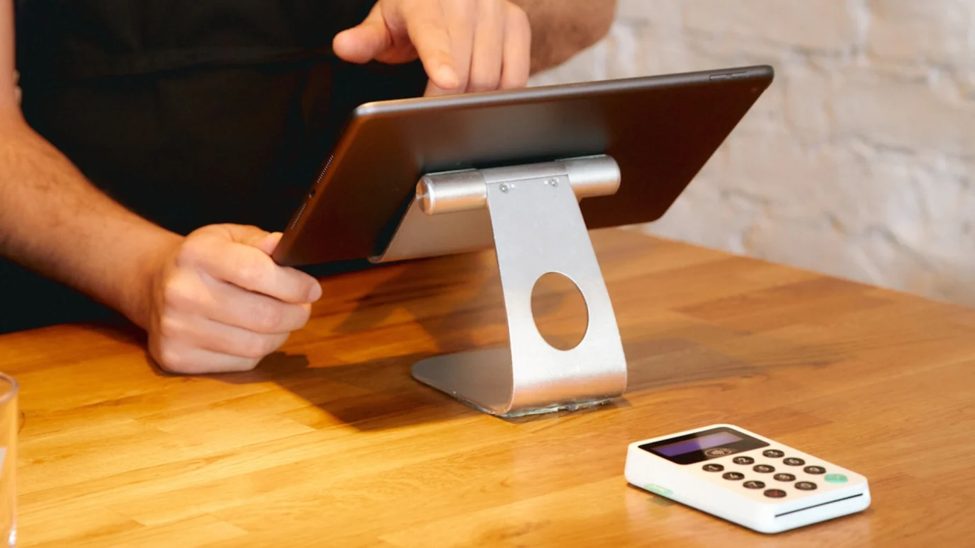 Close-up of a person using a tablet on a sleek metal stand next to a contactless payment terminal, emphasizing physical retail integration and streamlined operations for an accelerated startup.