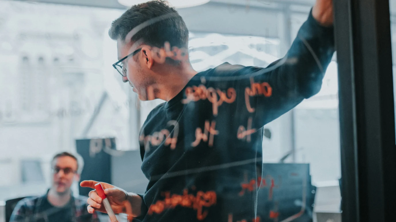 Strategy and discovery workshop: Product manager or designer writing notes on a clear glass whiteboard during a brainstorming or development meeting in a modern office.