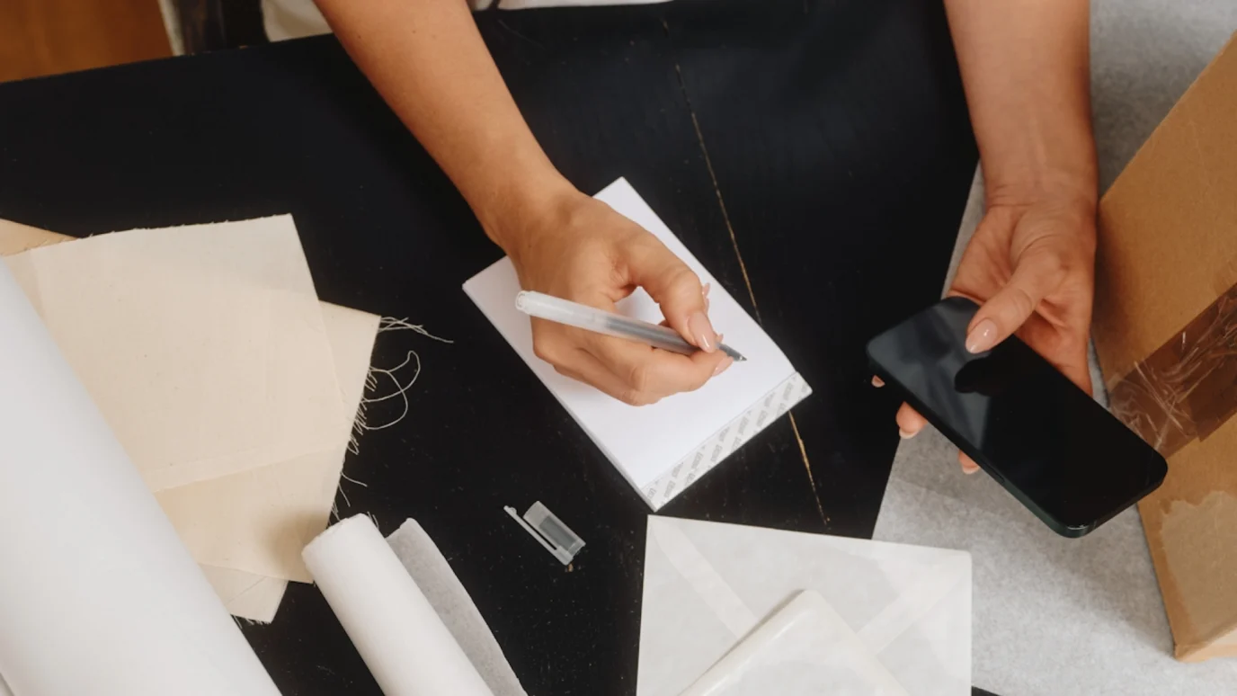 E-commerce strategy and planning: Close-up of hands writing notes in a small white notebook while holding a black smartphone, surrounded by packaging materials and a cardboard box.