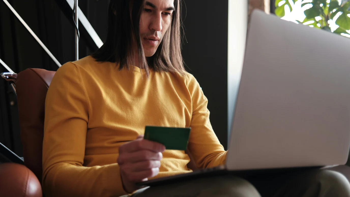 Online shopping or e-commerce transaction: Young man with long dark hair holding a green credit card while looking at a silver laptop, representing digital commerce and secure payment.