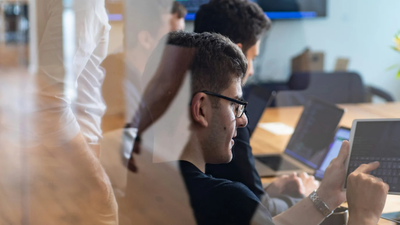 Group of people working in an office, reviewing code or data on tablets and laptops.