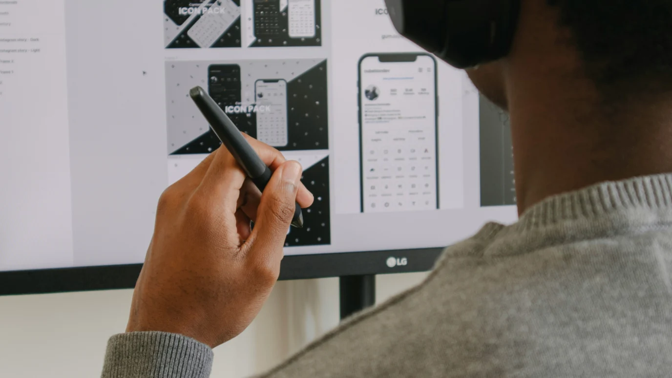 Designer holding a stylus, reviewing mobile UI designs on a computer monitor.