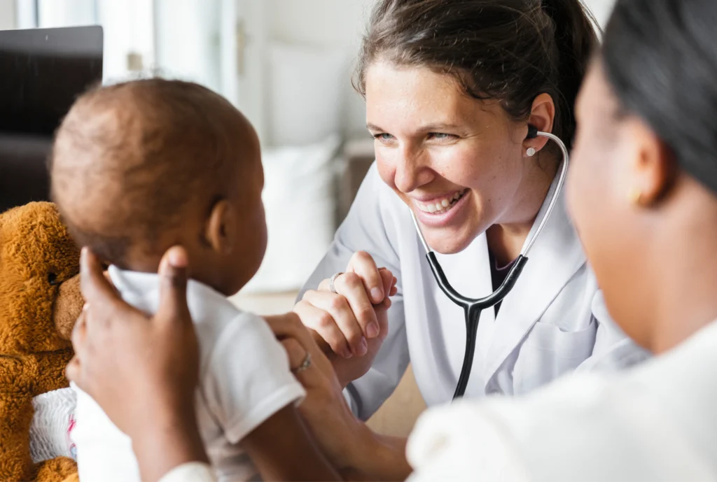 A female pediatrician wearing a stethoscope smiles while holding hands with an infant, symbolizing compassionate, high-quality pediatric care and family wellness.