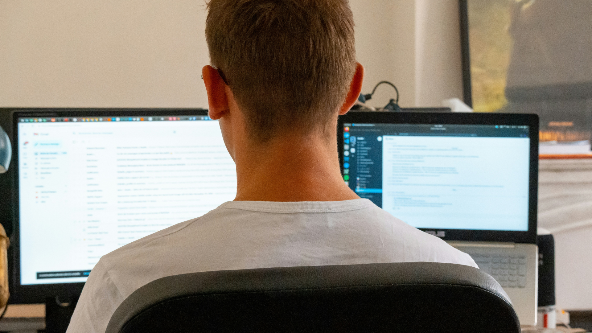 A person from behind sitting at a desk and working on a dual-monitor setup with a large computer screen and a laptop.